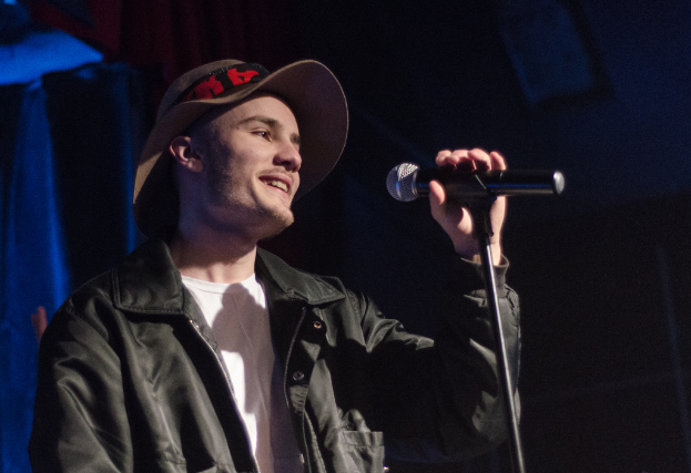 Ein Mann mit Hut und schwarzer Jacke, der fröhlich in ein Mikrofon spricht.