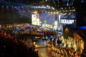 Menge auf einer hell beleuchteten Bühne mit Musikern, die Instrumente spielen, und einem großen Bildschirm im Hintergrund, der auf ein bevorstehendes Ereignis hinweist.