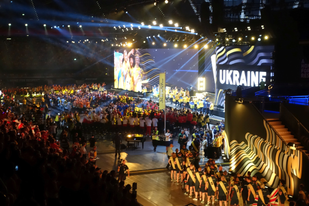 Menge auf einer hell beleuchteten Bühne mit Musikern, die Instrumente spielen, und einem großen Bildschirm im Hintergrund, der auf ein bevorstehendes Ereignis hinweist.