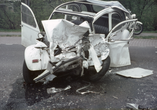 Ein Auto mit schwerer Frontschaden steht am Straßenrand, mit Bäumen, einem Gebäude und einem weiteren Fahrzeug im Hintergrund.