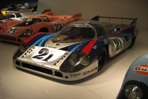 Ein Porsche 917K Le Mans Rennauto wird in einem Museum ausgestellt, umgeben von anderen Sportwagen und Menschen, mit einer Wand und einem Papier mit Text auf der linken Seite.