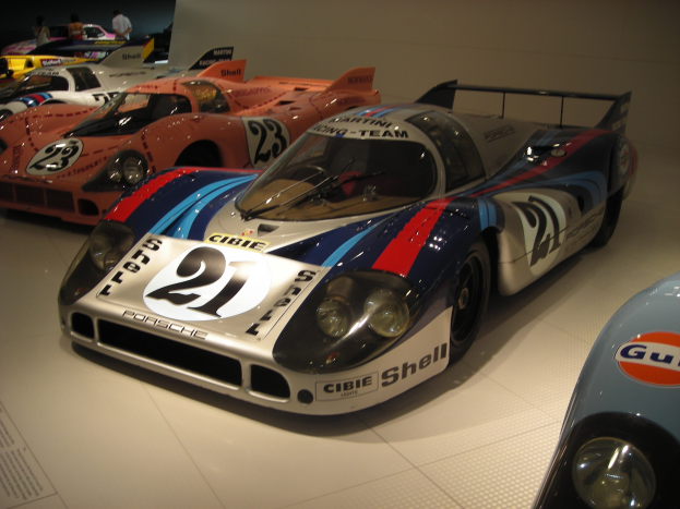 Ein Porsche 917K Le Mans Rennauto wird in einem Museum ausgestellt, umgeben von anderen Sportwagen und Menschen, mit einer Wand und einem Papier mit Text auf der linken Seite.