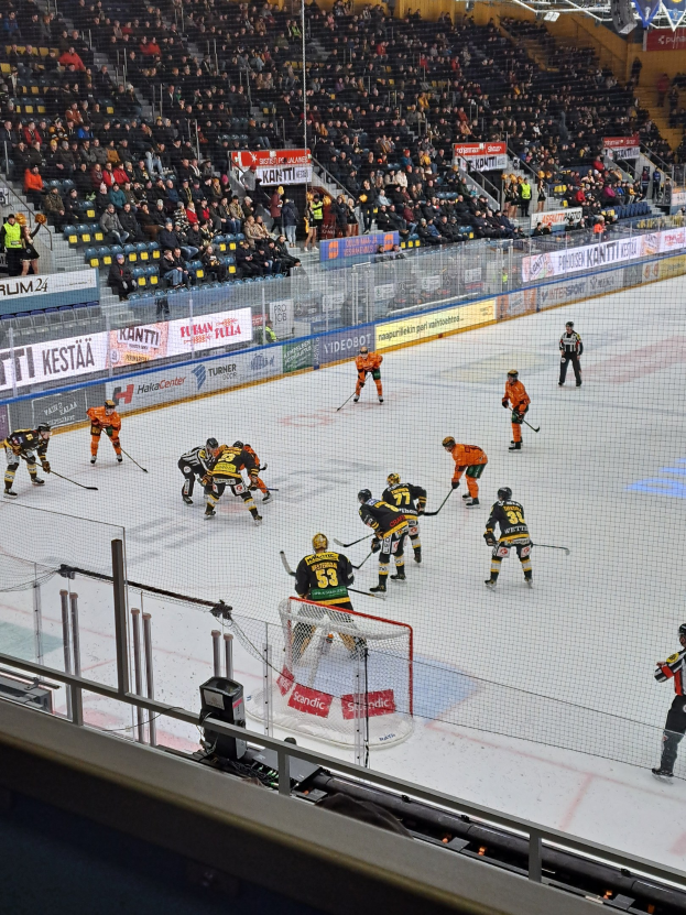 Eine Gruppe von Menschen, die Hockey auf einem Eisstadion spielen, mit einem Netz, einer Umzäunung, sitzenden Zuschauern, Bannern und Scheinwerfern.