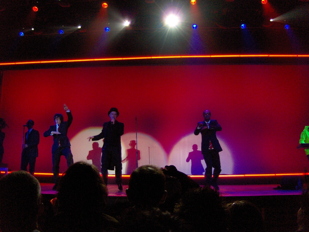 Ein Mann in einem schwarzen Kleid hält ein Mikrofon auf der Bühne, drei Personen tanzen neben ihm, während ein anderer Mann rechts am Piano spielt, ein Kameramann filmt die Szene und das Publikum schaut von der Seite zu, während Scheinwerfer von oben fokussiert sind.