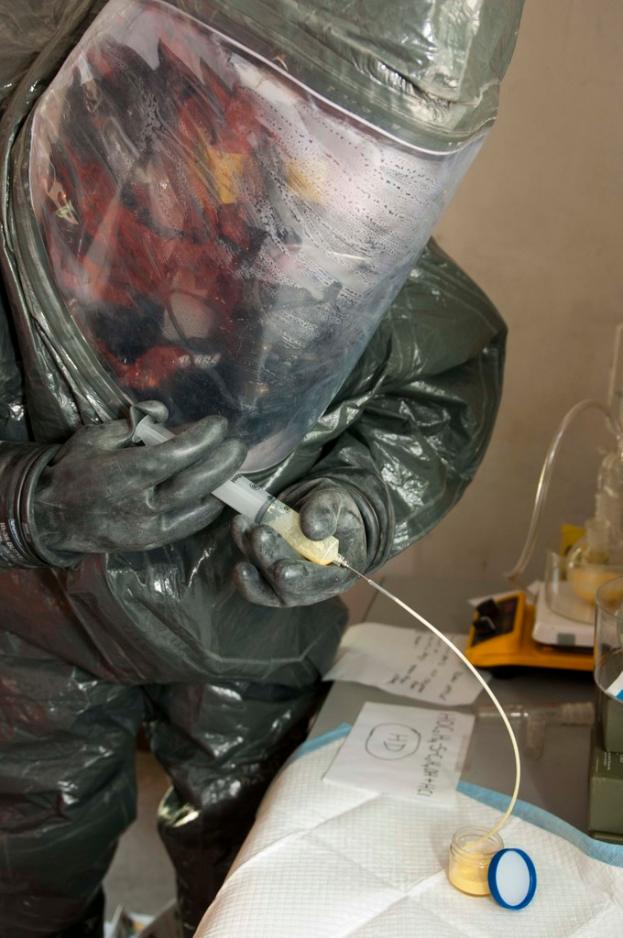 Eine Person in einem Laborkittel, Helm und Handschuhen injiziert eine Substanz in eine Flasche auf einem Tisch, der auch Papiere und eine Waage enthält.