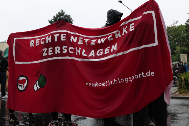 Eine Gruppe von Menschen steht auf einer Straße und hält ein rotes Banner hoch, auf dem "Rechte Netzwerke Zerschlagen" steht, mit Bäumen, Gebäuden und Laternen im Hintergrund, eine Person fährt rechts auf einem Fahrrad und eine weitere Person hält links einen Schirm unter einem klaren blauen Himmel.