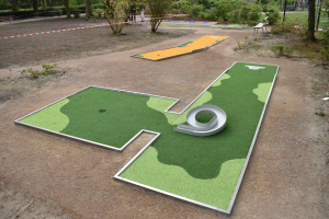 Minigolf-Platz in einem Park mit Grünfläche, Menschen in der Nähe, Gebäuden im Hintergrund und einem klaren blauen Himmel.