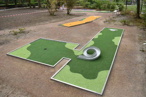 Minigolf-Platz in einem Park mit Grünfläche, Menschen in der Nähe, Gebäuden im Hintergrund und einem klaren blauen Himmel.