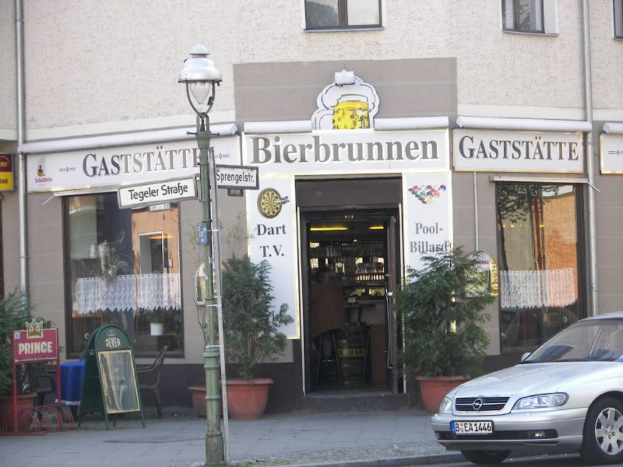 Ein Auto fährt auf der rechten Seite einer Straße, flankiert von einem Gebäude mit Pflanzen im Hintergrund und Stühlen auf der linken Seite, während eine Straßenlaterne in der Mitte steht.