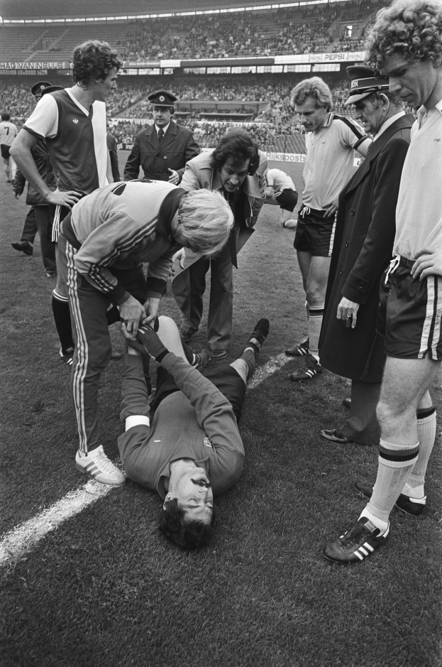 Schwarzes und weißes Bild eines Fußballspielers, der auf dem Boden liegt und von einer Gruppe von Männern umgeben ist, mit einer Menge im Stadionhintergrund.