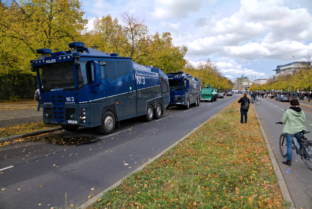 Eine Gruppe von Polizeifahrzeugen steht am Straßenrand, mit einer Person, die auf der rechten Seite Fahrrad fährt, viele Menschen halten Fahrräder, Gras und trockene Blätter auf dem Boden, Bäume, Gebäude und ein bewölkter Himmel im Hintergrund.