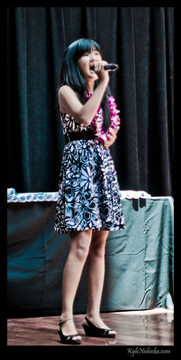 Eine Frau in einem bunten Kleid singt in ein Mikrofon auf der Bühne, mit einem Tisch voller Papiere und einem Vorhang im Hintergrund.