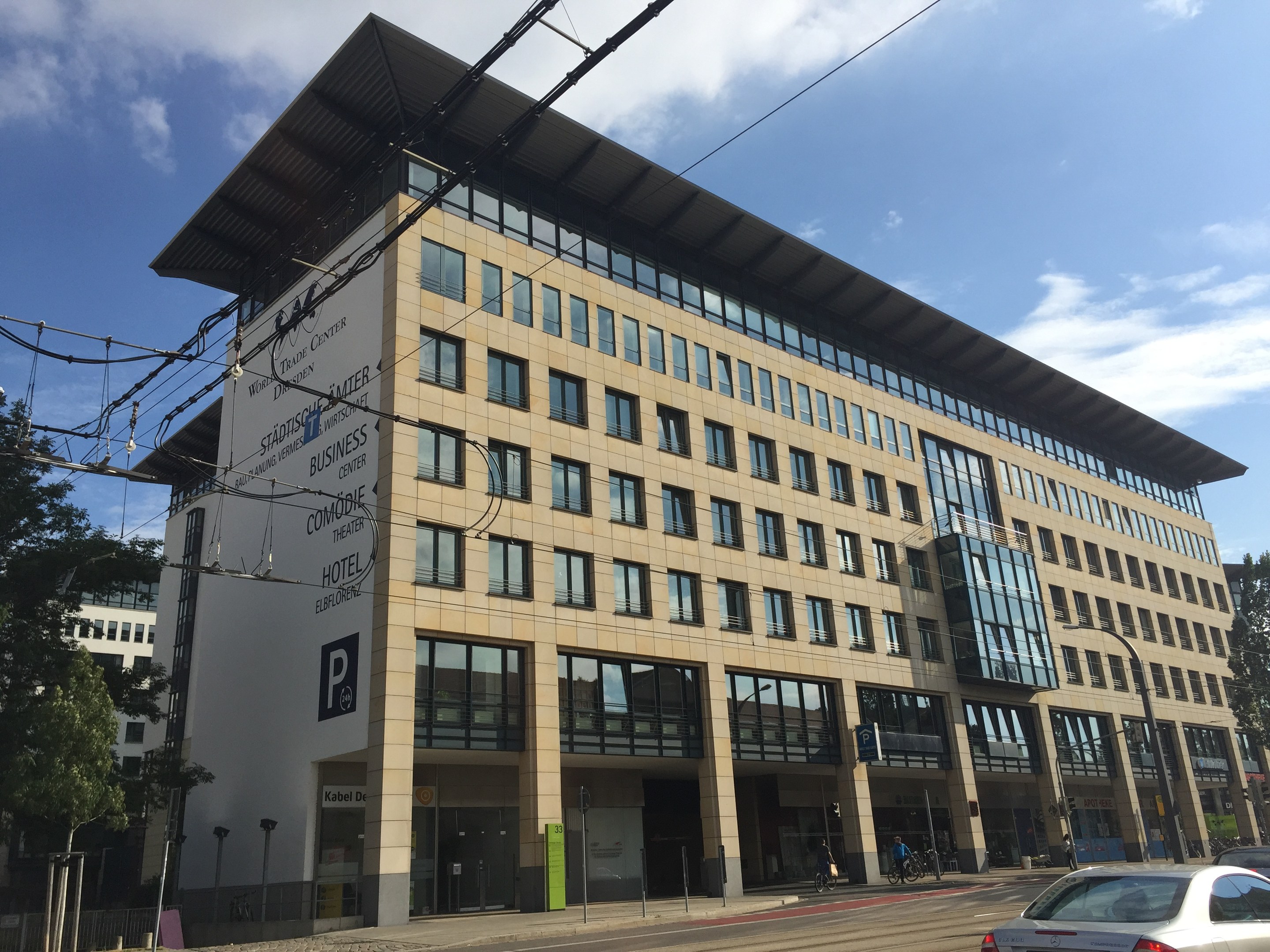 Großes modernes Gebäude mit zahlreichen Fenstern, beschriftet als Stadthalle Center für Geschäftsleute, umgeben von Bäumen und städtischer Infrastruktur einschließlich Straßenlaternen, Lichtern und Fahrzeugen, mit Fußgängern auf dem Bürgersteig unter einem bewölkten Himmel.