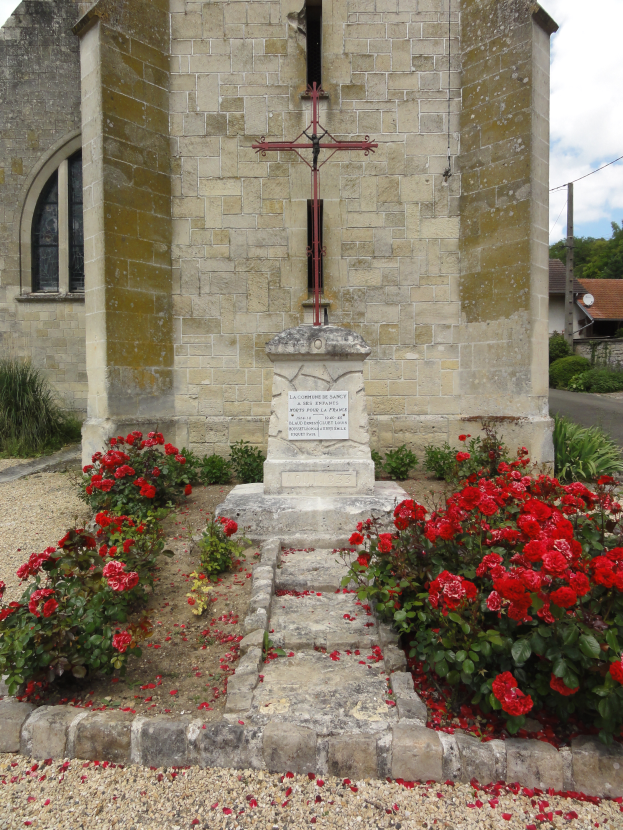 Ein steinernes Kreuz steht vor einer Kirche mit roten Rosen im Vordergrund, umgeben von Pflanzen und Bäumen, unter einem bewölkten Himmel.
