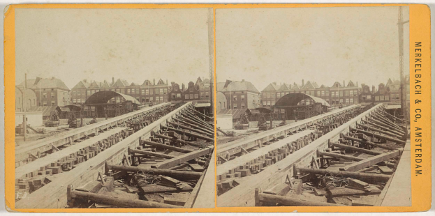 Ein altes Schwarz-Weiß-Foto einer Bahntrasse mit Gebäuden im Hintergrund, Text auf der rechten Seite mit der Aufschrift "Merkelbach & Co. Amsterdam" und verstreute hölzerne Gegenstände im Vordergrund.
