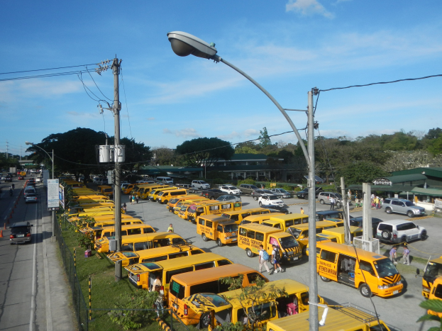 Eine lange Reihe gelber Schulbusse, die an einer Straße geparkt sind, mit Fußgängern auf dem Gehweg, Strommasten, Bäumen, Gebäuden und einem bewölkten Himmel im Hintergrund.