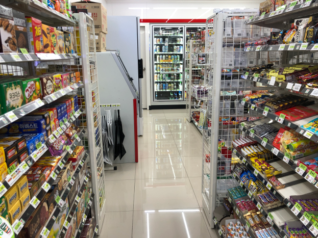 Ein Supermarktgang mit verschiedenen Lebensmittelarten auf Regalen, Preisetiketten sichtbar, ein Kühlschrank im Hintergrund und Deckenbeleuchtung.