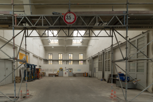 Großes Lagerinneres mit ausgedehnten Gerüsten, Verkehrszeichen, Metallstangen, einem Schild, Maschinen, einer Tür, Fenstern, Wänden, Deckenleuchten und einem Dach.