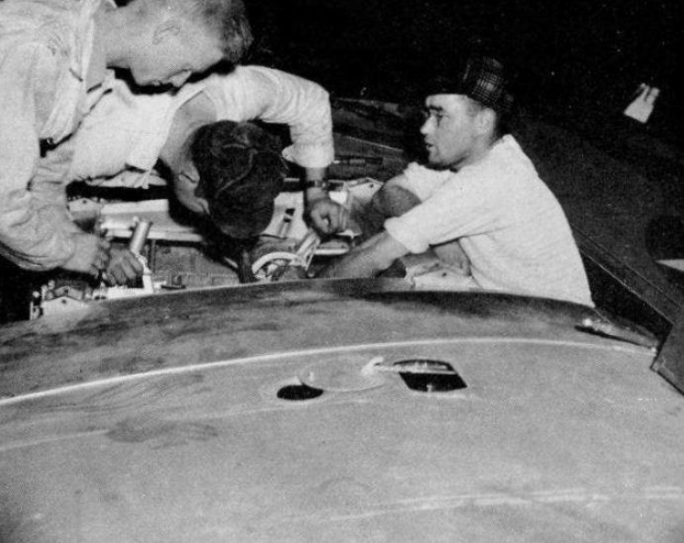 Drei Männer in Baseballkappen reparieren ein schwarz-weißes Auto in einer Garage, umgeben von Werkzeugen und Materialien.