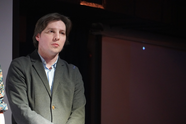 Zwei Personen, ein Mann im Anzug und eine Frau, stehen Seite an Seite auf einer Bühne mit ernsten Gesichtern.