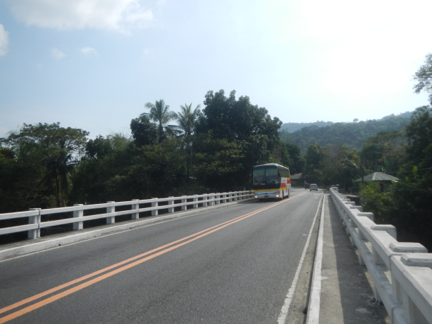 Ein Bus fährt über eine Brücke mit Geländern auf beiden Seiten, umgeben von Bäumen und einem bewölkten Himmel.
