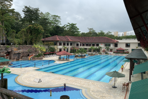 Großes Schwimmbad im Ritz-Carlton, Kota Kinabalu, mit Menschen, Sonnenschirmen, Stühlen, Tischen, Pflanzen, Bäumen, Felsen und Gebäuden, einschließlich eines Schuppens auf der rechten Seite und Himmel im Hintergrund.