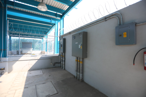 Ein Raum mit einem Feuerlöscher auf der rechten Seite, elektrischen Schaltern und Rohren an der Wand, einer Deckenlampe und einem Zaun im Hintergrund, was auf eine elektrische Umspannstation hinweist.