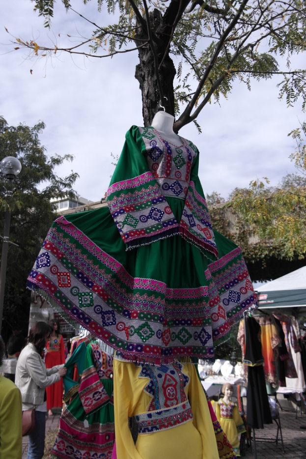 Eine Schaufensterpuppe steht inmitten eines belebten Einkaufsbereichs mit mehreren Geschäften, einer Person, die Kleider an einer Stange arrangiert, einem Gebäude und Bäumen im Hintergrund.