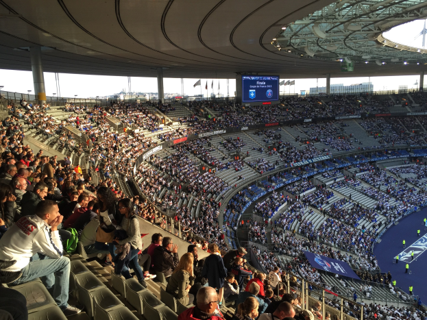 Große Zuschauermenge in einem Stadion bei einem Fußballspiel mit einer Bühne, Flaggen, Stangen und einem Bildschirm im Hintergrund.