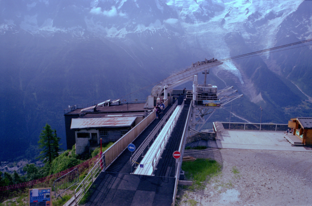 Seilbahn fährt einen schneebedeckten Berg hinauf, mit einem Hintergrund aus schneebedeckten Bergen, die über eine Straße mit Schildern, Geländern, Vegetation und Häusern fahren, mit ein paar Menschen sichtbar.