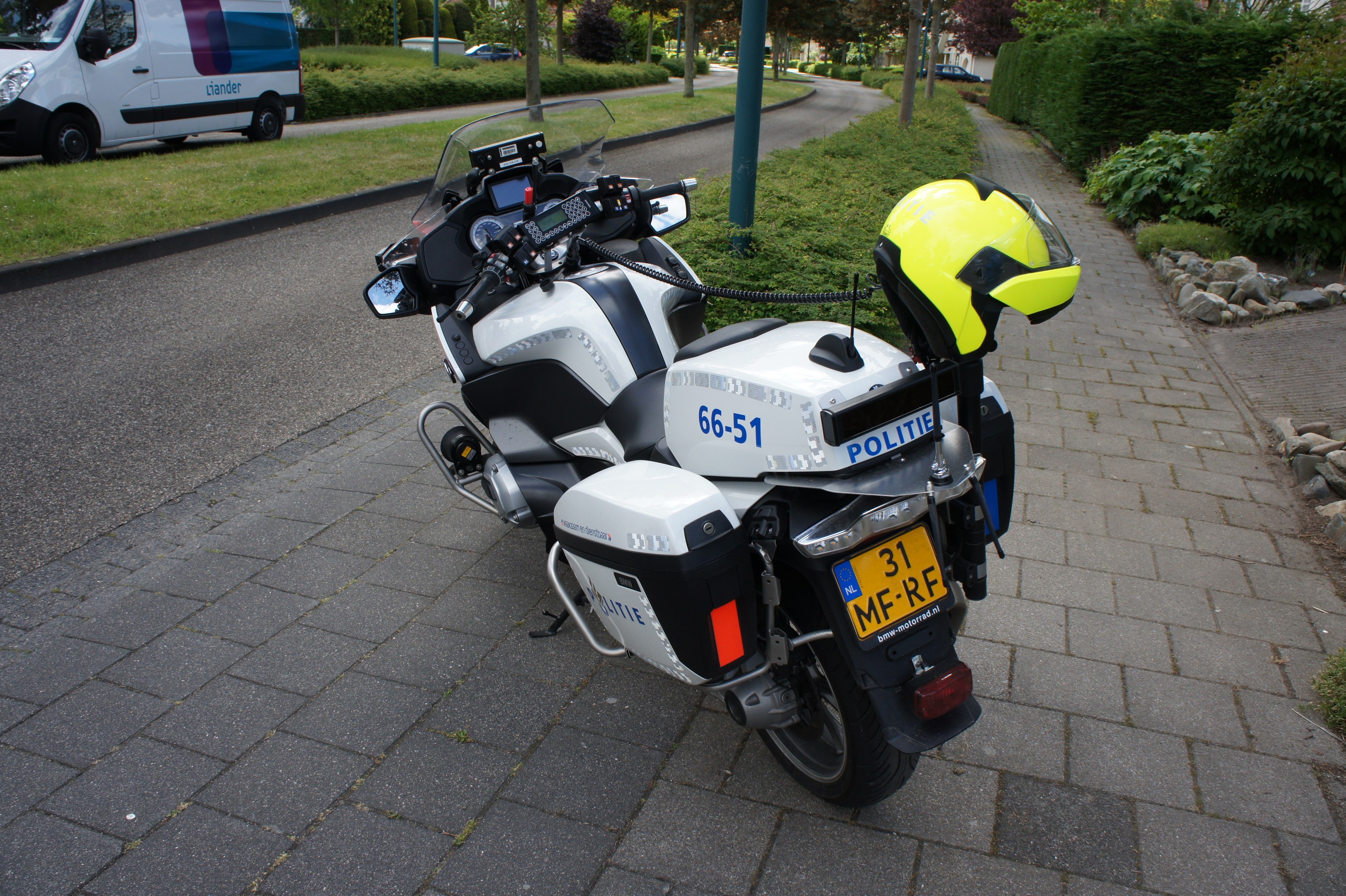Ein polizeiliches Motorrad am Straßenrand geparkt mit einem Helm darauf, umgeben von Bäumen, Gebäuden und anderen Fahrzeugen.