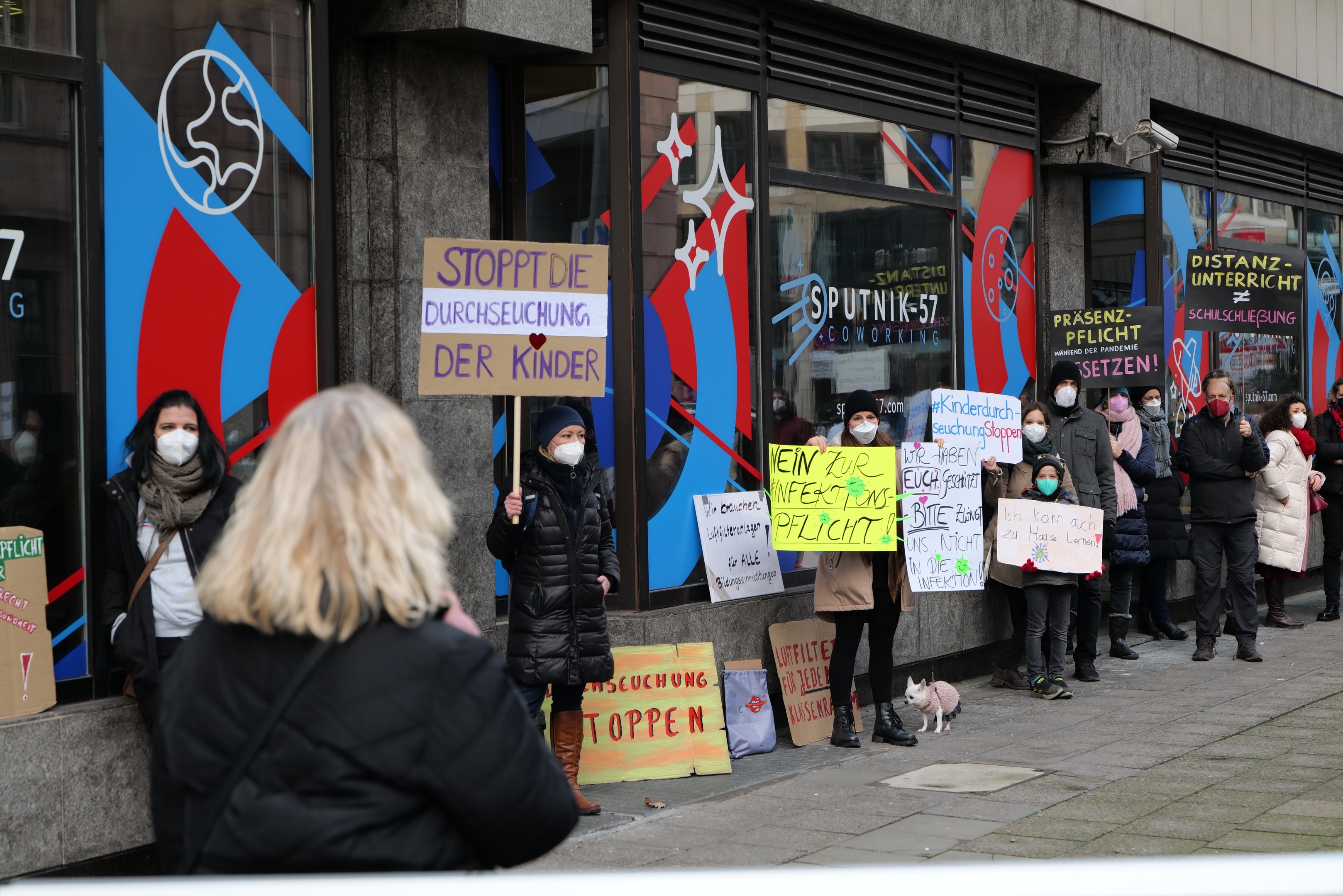 Eine Gruppe von Menschen mit Masken und Schildern protestiert vor einem Gebäude mit Glaswänden und -türen, begleitet von Tafeln mit Text und Logos, Kameras und einem Hund.