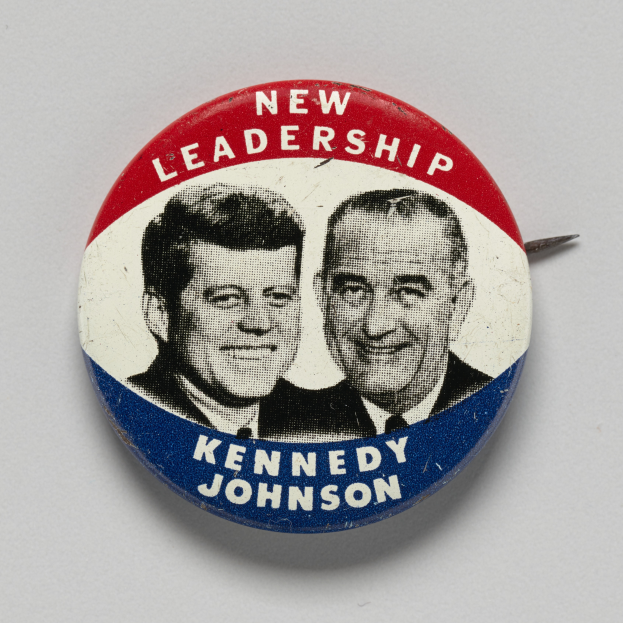 Ein Ansteckbutton auf einer weißen Oberfläche zeigt zwei Fotos von Kennedy und Johnson mit dem Text "Neue Führung" darunter.