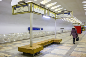 Eine Frau in einem Mantel trägt eine Tasche, während sie durch eine belebte Moskauer U-Bahn-Station mit Bänken, beleuchteten Tafeln und anderen Einrichtungsgegenständen geht.