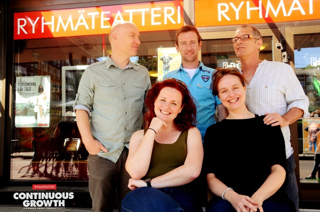 Drei Männer stehen und zwei Frauen sitzen vor ihnen, alle lächeln, mit Geschäften im Hintergrund und einer Tafel mit der Aufschrift "RHYME TEATTERI" über einem der Geschäfte.