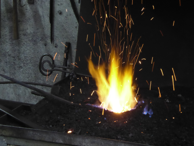 Ein Schmied arbeitet an einem Metallstück, Funken fliegen, mit Metallstangen und einer Wand links und einem Feuer rechts.