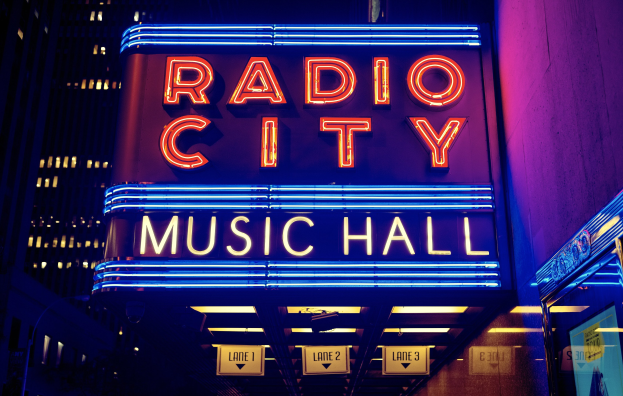 Hell erleuchtetes Radio City Music Hall in New York City mit hohen umliegenden Gebäuden, das eine 'Radio City'-Leuchtschrift und zusätzliche Schilder auf der rechten Seite zeigt.