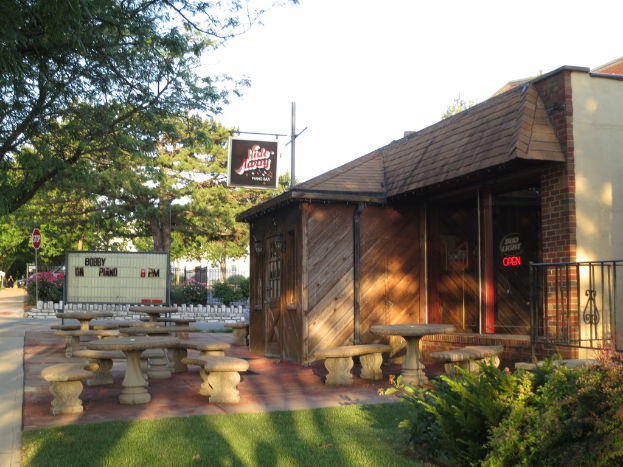 Ein Haus mit einem Namensschild, Bänken, Gras, Pflanzen, einer Straße, einem Container, einem Schild, Bäumen und einem Himmel.