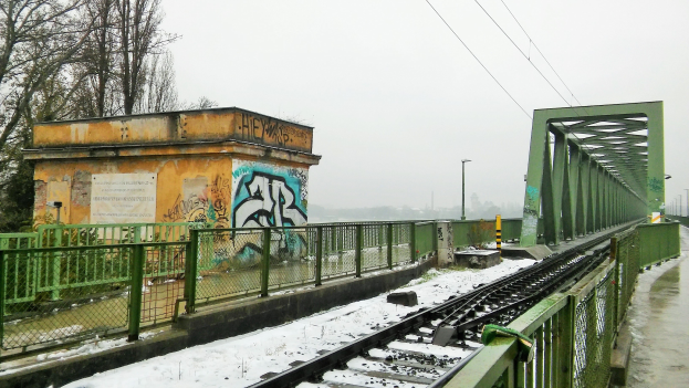 Eisenbahnschiene mit Graffiti an der Seite, umgeben von Gelöndern, Pfosten, Lichtern, Drähten und Bäumen, mit Schnee bedeckt und mit einem sichtbaren Himmel im Hintergrund.