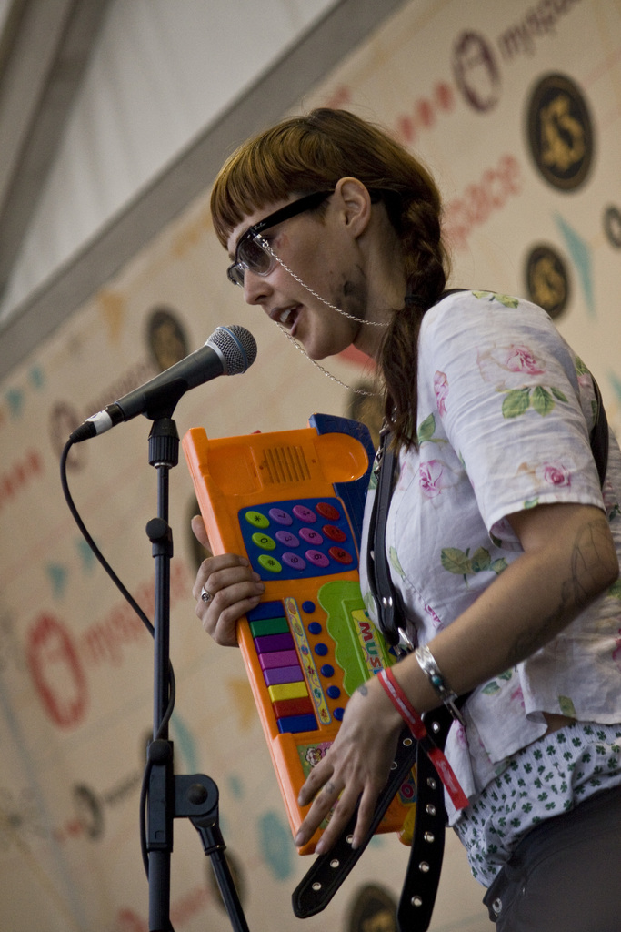 Eine Frau in einem weißen Hemd und Sonnenbrille singt in ein Mikrofon und spielt Klavier, mit einem Plakat im Hintergrund.