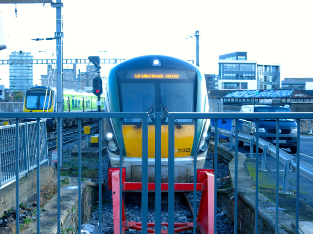 Zwei Züge auf dem Gleis mit einem Zaun im Vordergrund, Pfosten auf dem Land, einer mit einer Signalleuchte, ein Auto auf der Straße rechts, Gebäude im Hintergrund und Himmel oben.