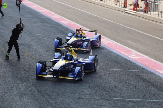 Zwei Formel-E-Autos auf einer Rennstrecke mit einem Fahne schwingenden Mann in der Nähe, während ein paar Menschen links gehen und andere im Hintergrund hinter Gittern sichtbar sind.