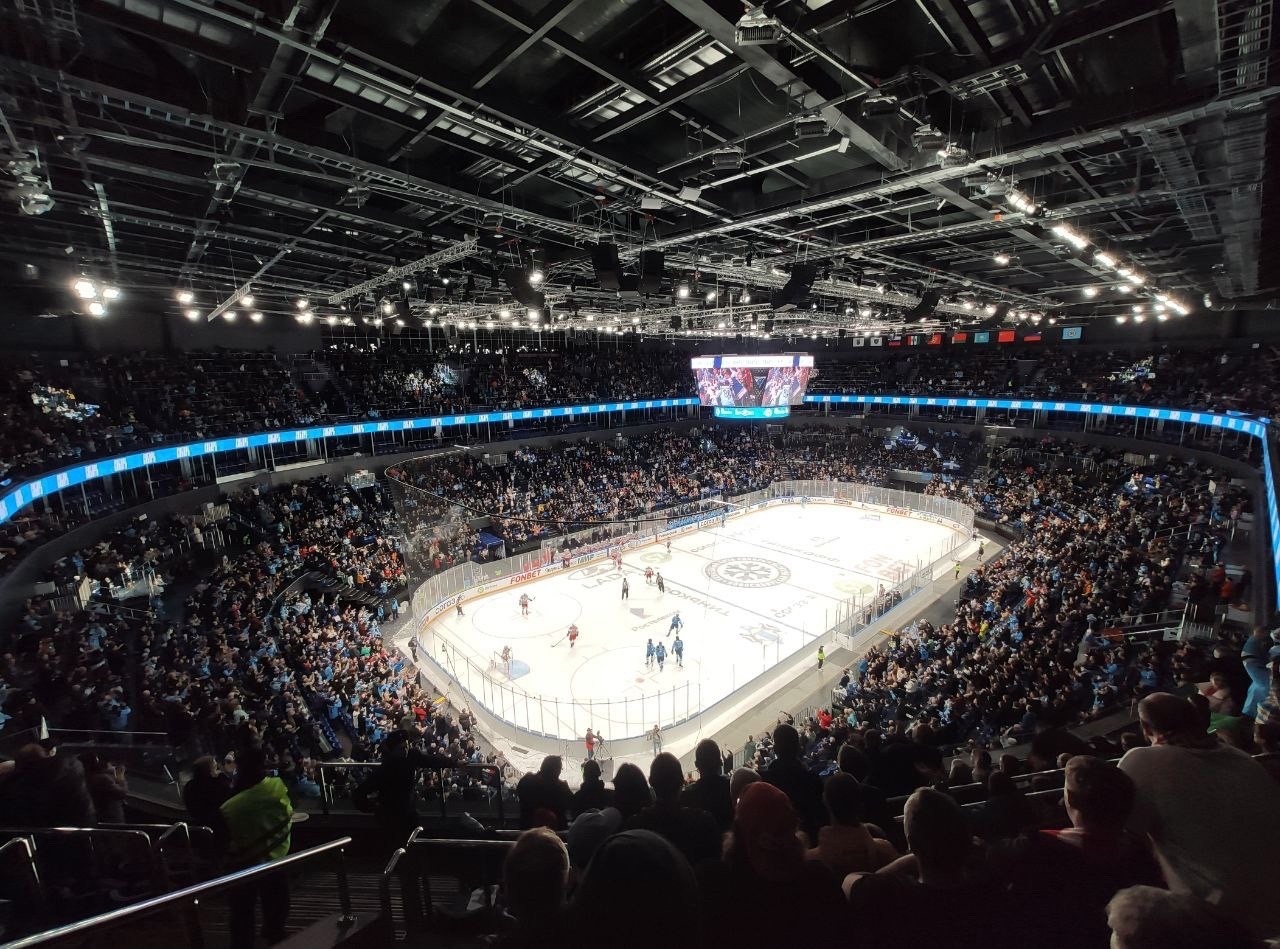 Ein Eishockeyspiel wird im O2 Arena in London gespielt, mit Zuschauern auf den Rängen und Spielern auf dem Eis, unter hellen Scheinwerfern mit einem Bildschirm an der Spitze des Bildes.