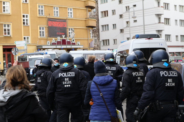 Polizeibeamte in Uniform stehen vor einer Menge von Helmträgern während einer Demonstration in Berlin, mit Fahrzeugen, Gebäuden und einem Banner im Hintergrund.