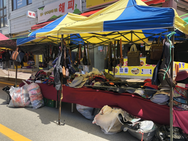 Ein Markt mit Zelten, die Taschen, Hüte und andere Gegenstände präsentieren, vor Gebäuden mit Fenstern und Texttafeln.