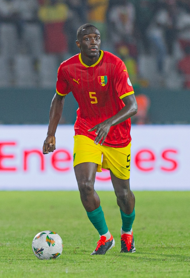 Ein Mann in rot-gelber Uniform spielt Fußball auf einem Feld, trägt Turnschuhe und hat eine Tafel im Hintergrund und einige Zuschauer auf den Rängen.