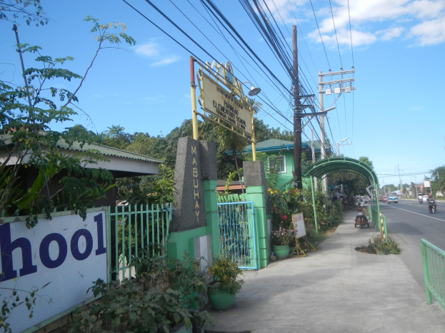 Schuleingangsschild an der Seite einer Straße mit vorbeifahrenden Fahrzeugen, einem Torbogen, einem Tor, einer Namenstafel, umgeben von Grün, Strommasten mit Drähten und einem bewölkten Himmel.