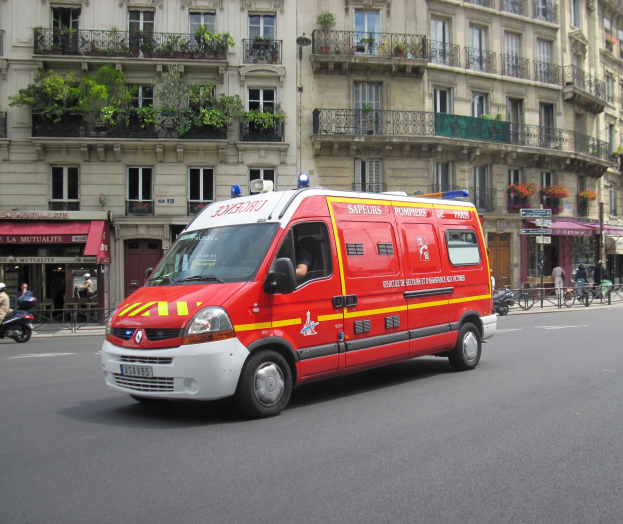 Rettungswagen auf der Seite der Straße vor einem Gebäude mit Fenstern, Geländern, Pflanzen und anderen Gegenständen geparkt, mit ein paar Menschen und Fahrrädern im Hintergrund.