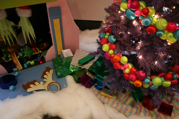 Drinnen steht rechts ein geschmückter Weihnachtsbaum mit Lichterkette und Schmuck, in der Mitte Geschenkboxen und Dekorationen, links oben eine Hütte mit Dekorationen und unten Baumwolle und Wolle.