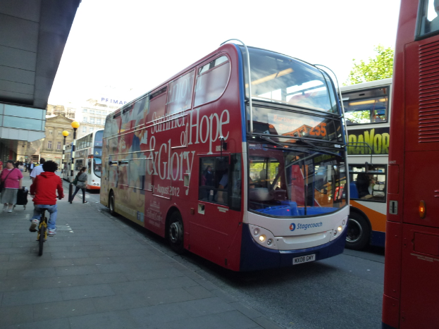 Eine Person fährt mit dem Fahrrad auf einem Gehweg mit vielen Menschen, Bussen mit Text, Gebäuden und Laternen im Hintergrund.
